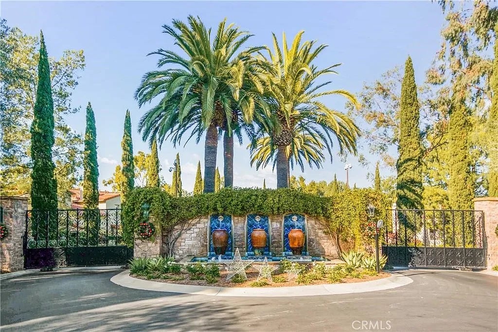 Gated property entrance with a three-basin fountain, stone wall, and tall palm trees.