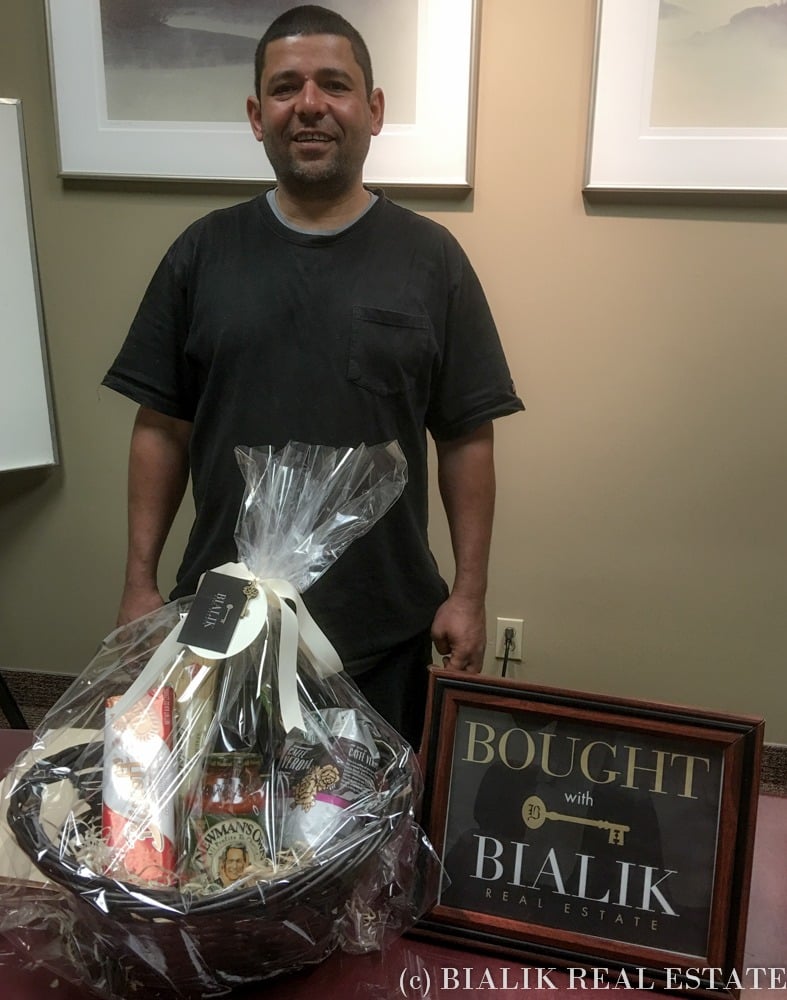 A smiling man stands with a wrapped gift basket and a framed Bialik Real Estate sign.