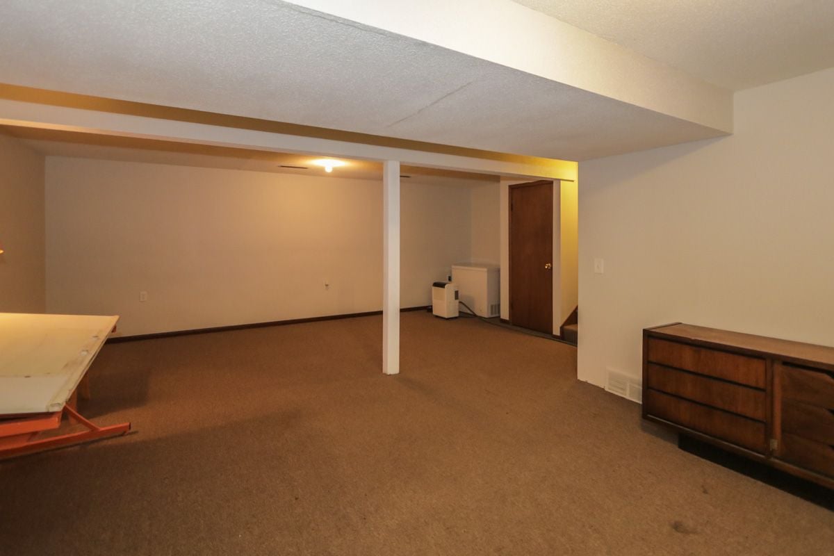 Carpeted basement room with a central support post, low ceiling beams, and sparse furniture.