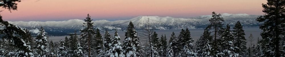 Snow-covered evergreens frame a lake and mountain range beneath a pink dusk sky.