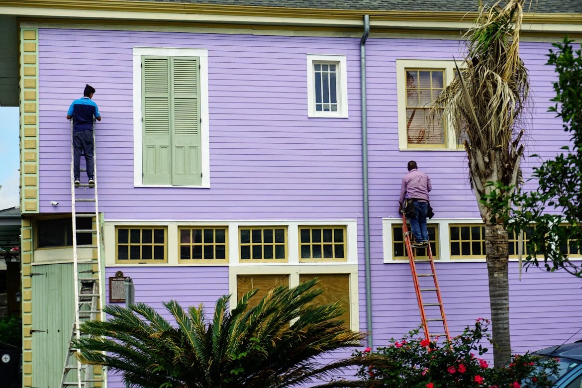 Two workers on ladders repairing and painting the exterior siding of a house.