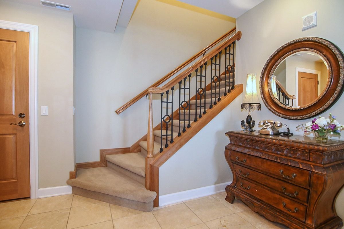 Foyer with a carpeted staircase, wood-and-iron railing, and carved chest beneath a round mirror.
