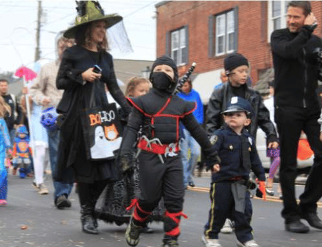 Children in ninja and police costumes walk in a Halloween parade beside an adult dressed as a witch.