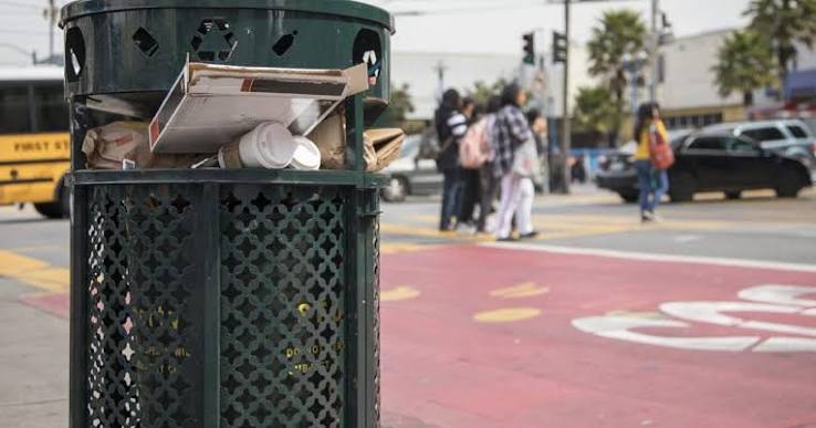 Public trash receptacle on urban sidewalk with pedestrians crossing near bike lane