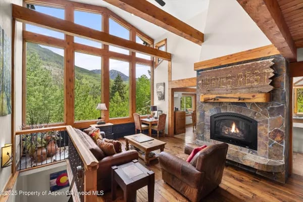 Living room with a stone fireplace, timber beams, and large windows overlooking forested mountains.