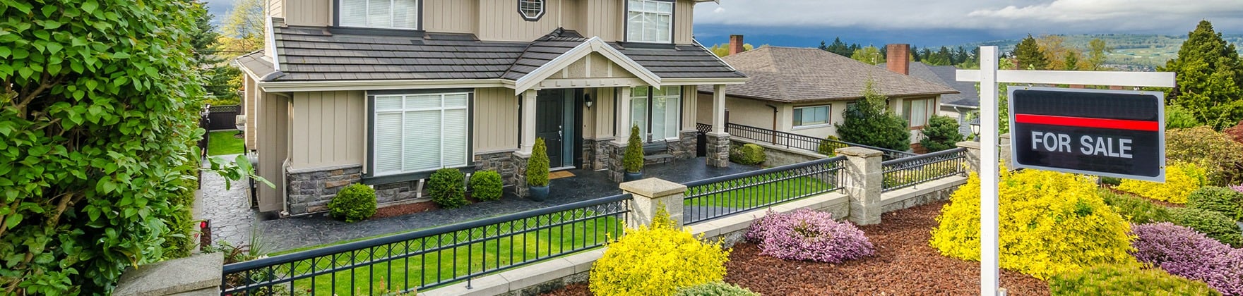 Two-story beige house with stone accents, landscaped garden, and a prominent FOR SALE sign.