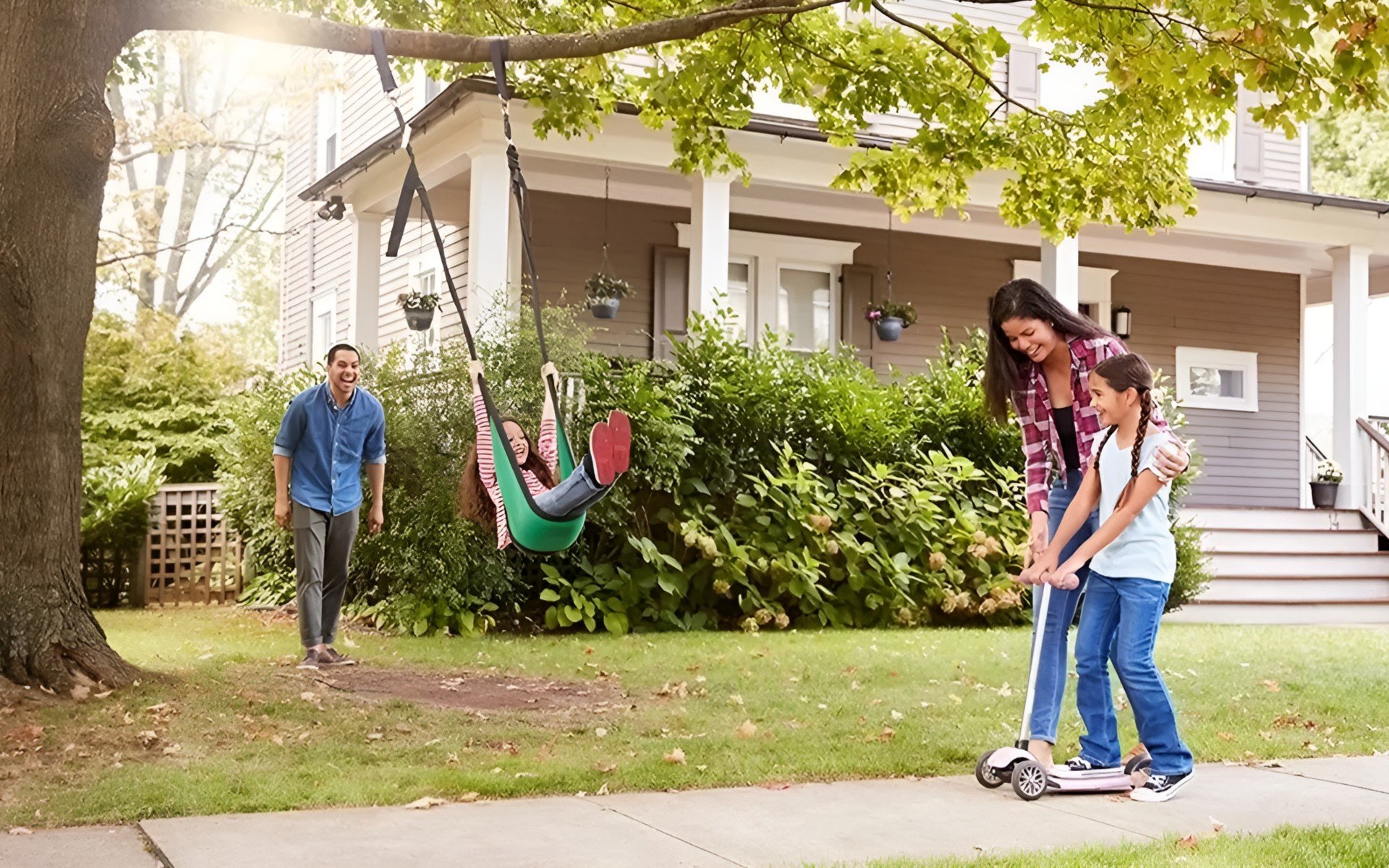 Family playing in a leafy front yard, with a child on a swing and another on a scooter.