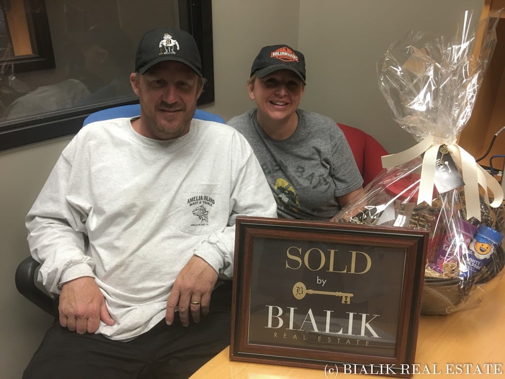 Two adults sit beside a framed Bialik Real Estate sold sign and a gift basket.