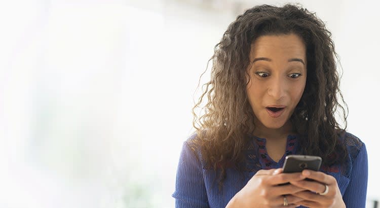 A woman looking at her phone, expressing surprise or excitement, captured in a moment of discovery or reaction to good news.