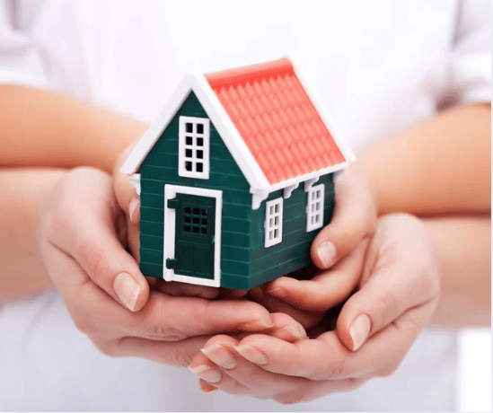 Hands cup a small green model house with white trim and a red roof.