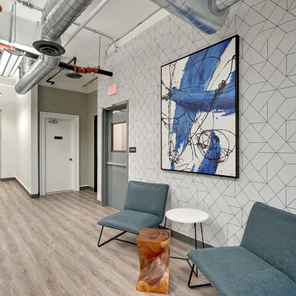 Building lobby with an elevator, blue lounge chairs, and large abstract artwork on a geometric wall.