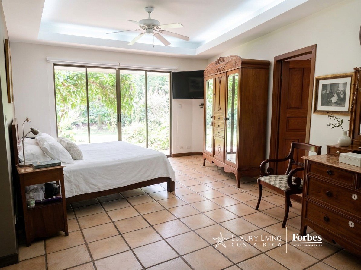 Furnished bedroom with a white bed, carved wooden wardrobe, and sliding glass doors overlooking greenery.