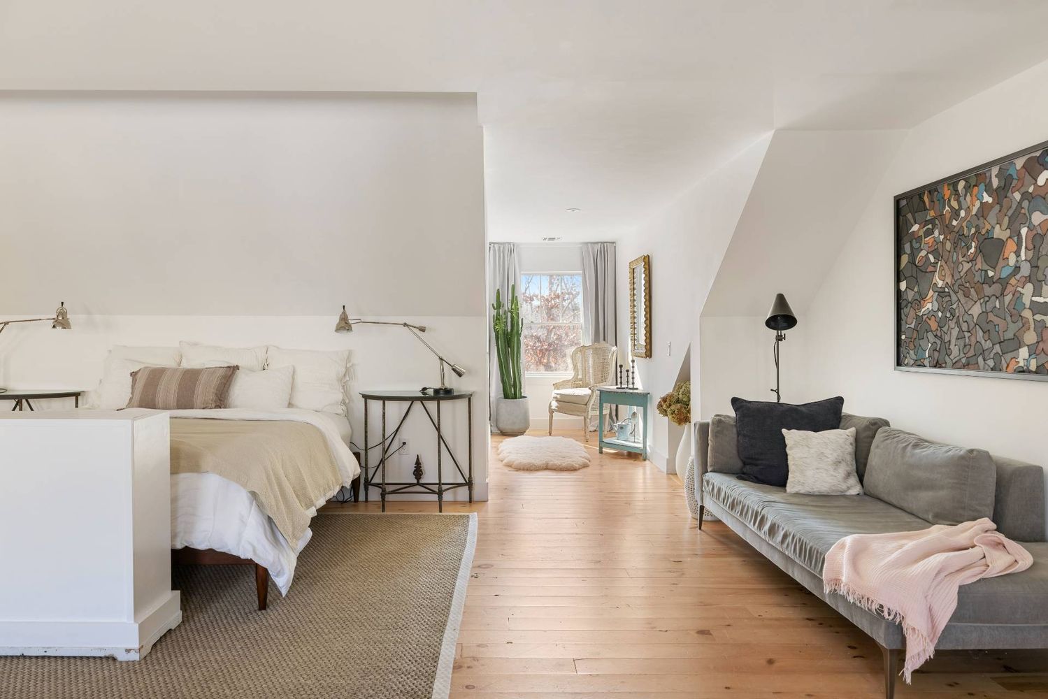 Bedroom with a white bed and adjoining gray sofa, light wood floors, and a tall cactus by the window.