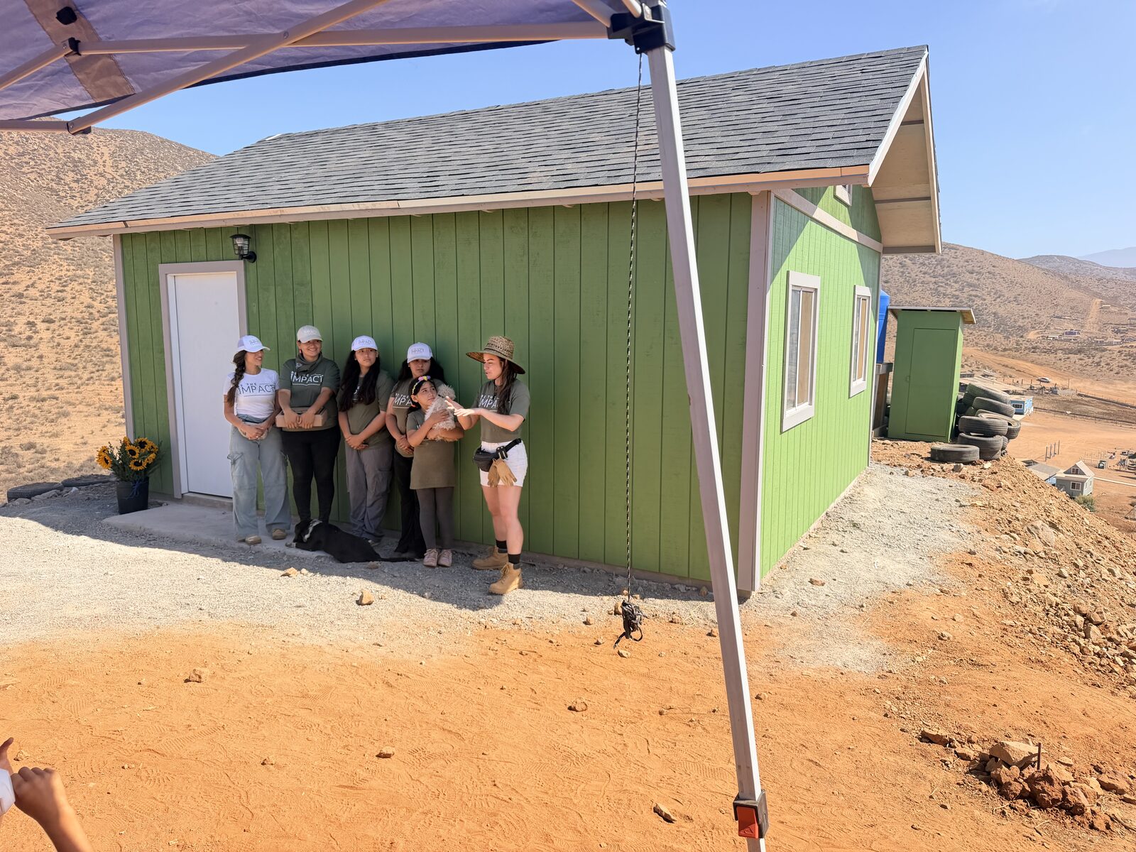 The completed green house with a shingle roof, white door and windows, and the family and volunteers standing in front of it