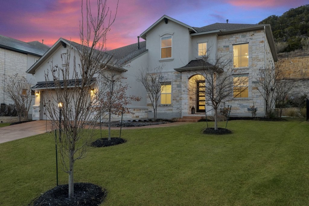 Two-story stone-and-stucco home with a broad lawn and warmly lit windows at dusk.
