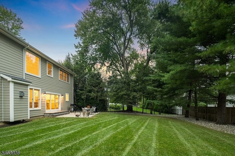 Rear of a gray-sided home with lit windows, patio furniture, and a broad striped lawn beneath mature trees.