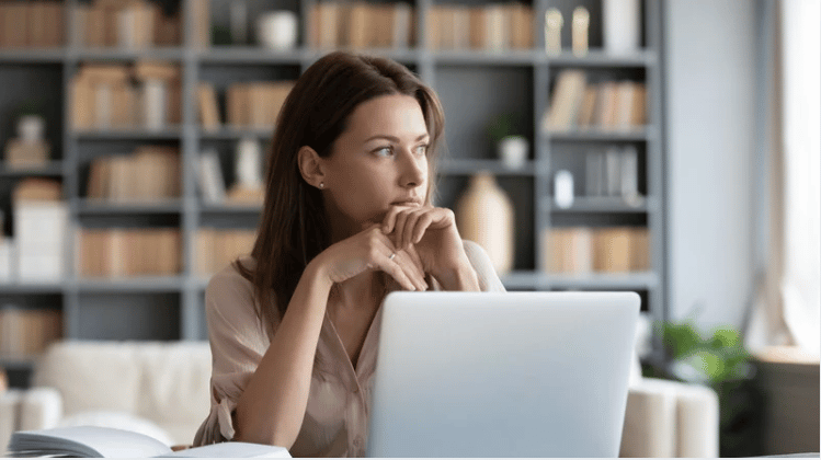 Thoughtful woman at a laptop, looking toward a window in a bright home office.