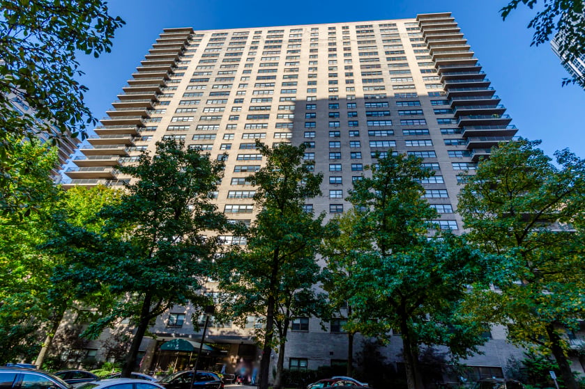 High-rise apartment building with projecting balconies, viewed from below through leafy trees.
