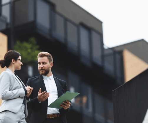 A man holding a green tablet talks with a woman outside a modern glass building.