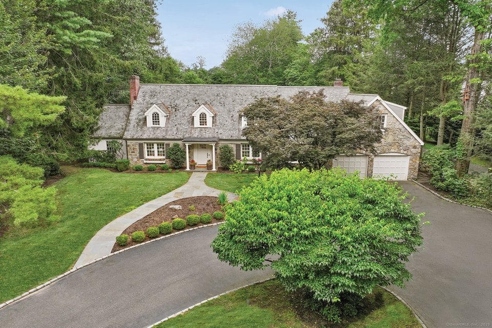 Stone-and-shingle home with slate roof, dormer windows, and attached two-car garage amid mature landscaping.