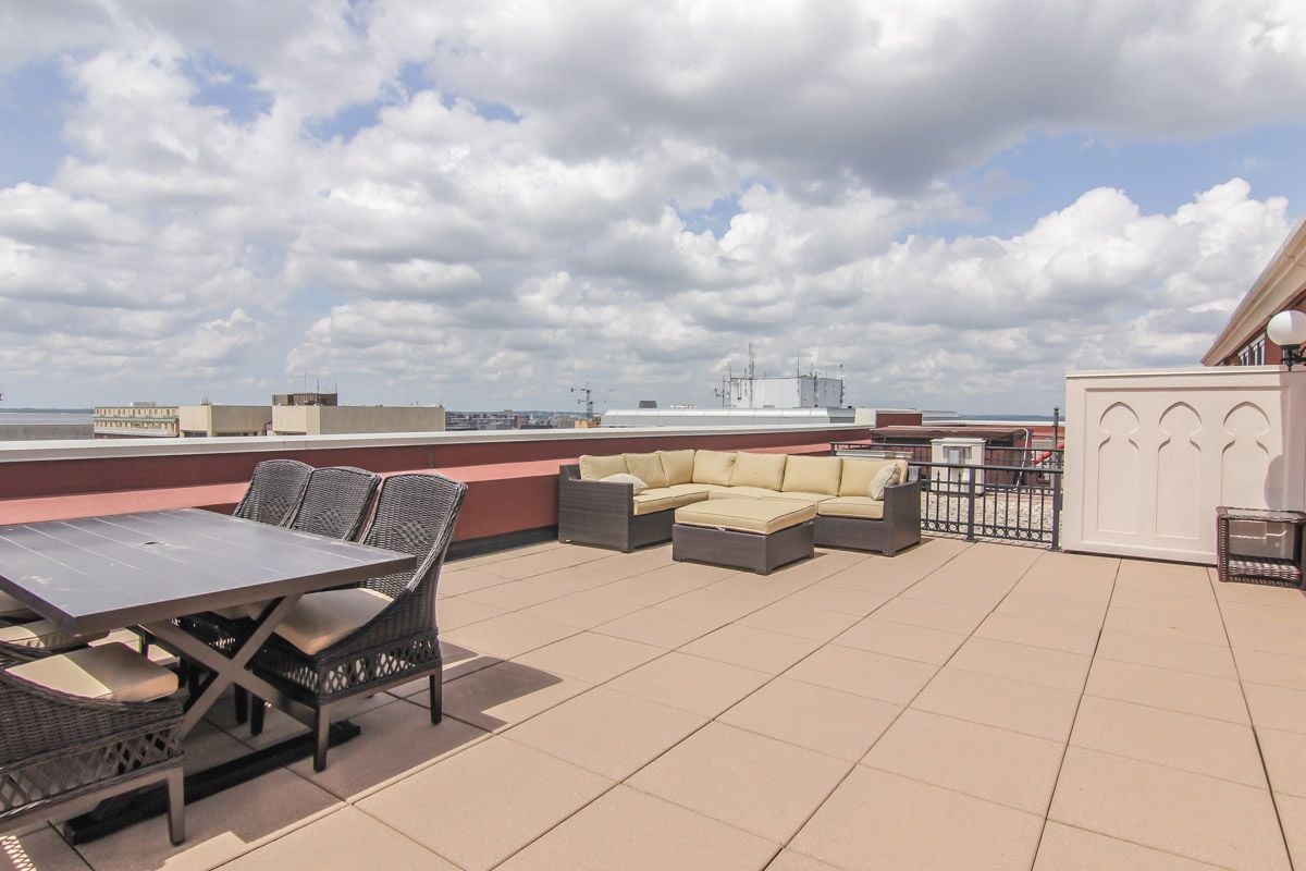 Furnished rooftop terrace with tiled flooring, wicker dining set, and sectional sofa overlooking distant buildings.