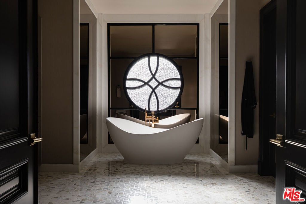 Bathroom with a freestanding tub beneath a large circular patterned window and pale mosaic tile floor.