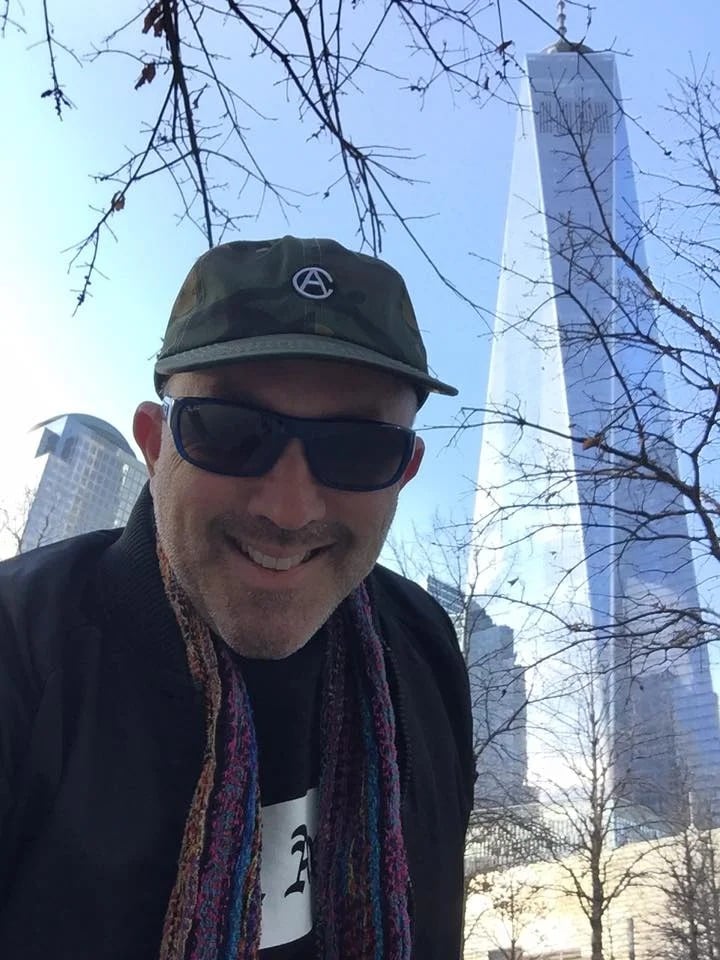 Man in sunglasses and a dark cap poses beneath One World Trade Center.