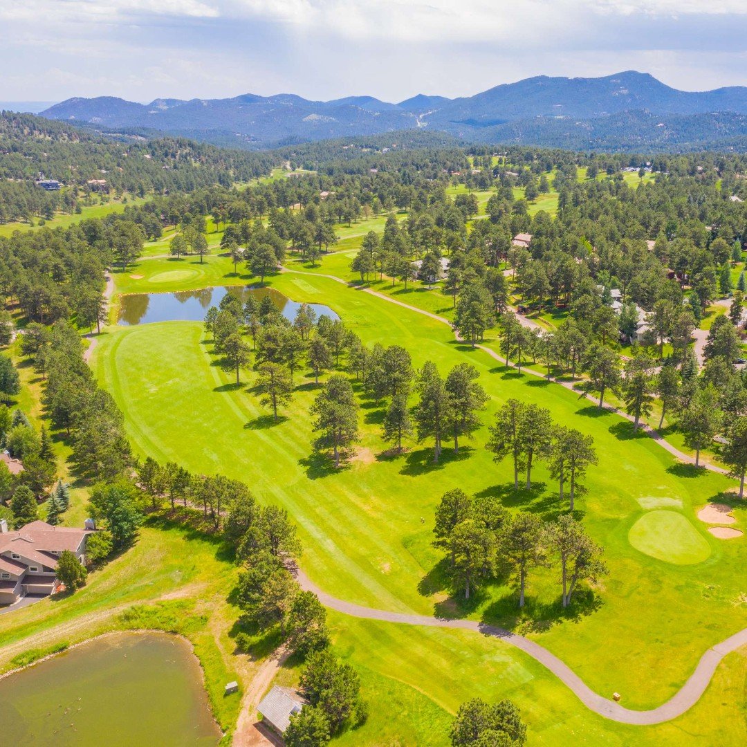 Aerial golf course with ponds and winding paths beneath forested Rocky Mountain ridges.