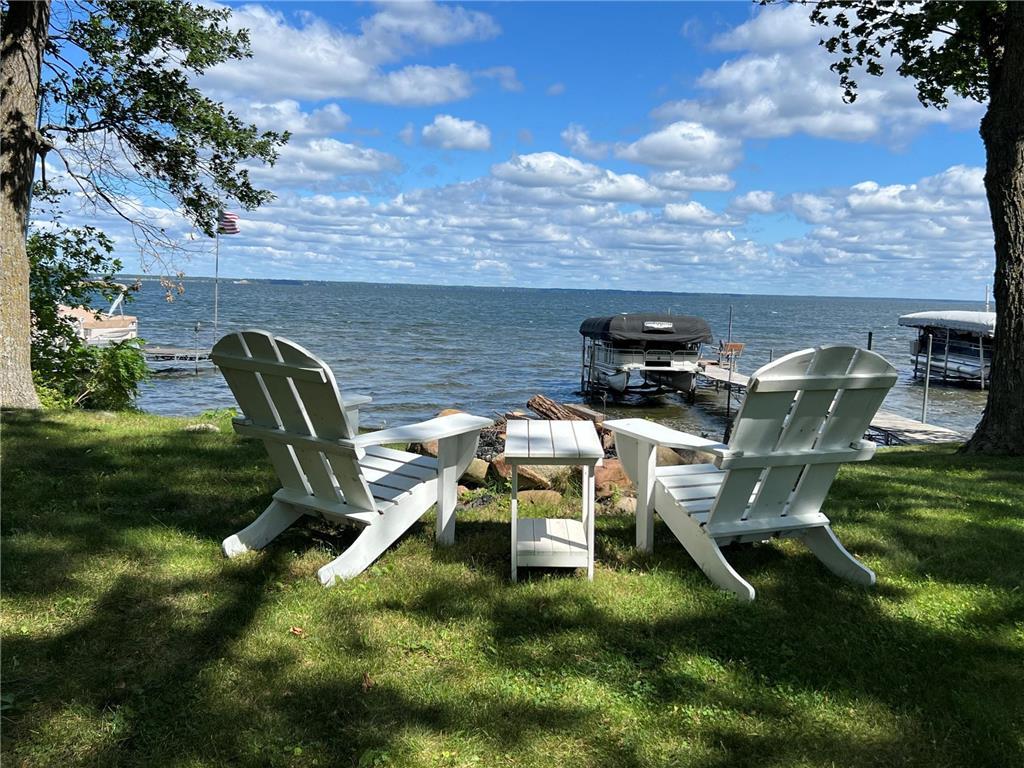 Two Adirondack chairs and a table face a lake from a shaded grassy shore with docks and boats.