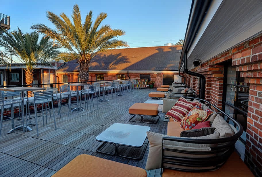 The image showcases Ulele's stunning outdoor terrace in Tampa Heights, featuring cozy lounge seating, modern high-top tables, and vibrant palm trees. This inviting space, surrounded by the restaurant's historic brick architecture, is perfect for enjoying a meal or drinks under the beautiful Florida sky.
