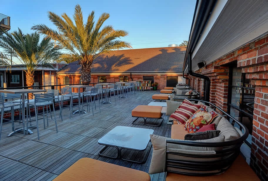 The image showcases Ulele's stunning outdoor terrace in Tampa Heights, featuring cozy lounge seating, modern high-top tables, and vibrant palm trees. This inviting space, surrounded by the restaurant's historic brick architecture, is perfect for enjoying a meal or drinks under the beautiful Florida sky.