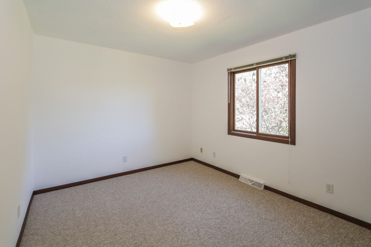 Empty bedroom with beige carpet, dark wood trim, and a window overlooking flowering trees.