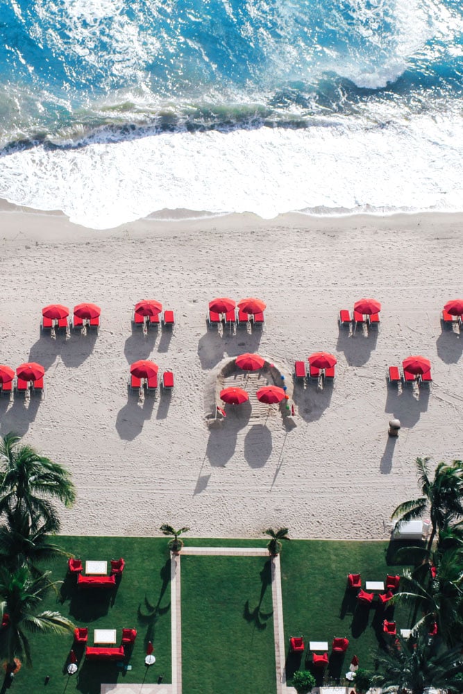Aerial resort beach with red umbrellas, foamy waves, and palm-fringed lawn seating.