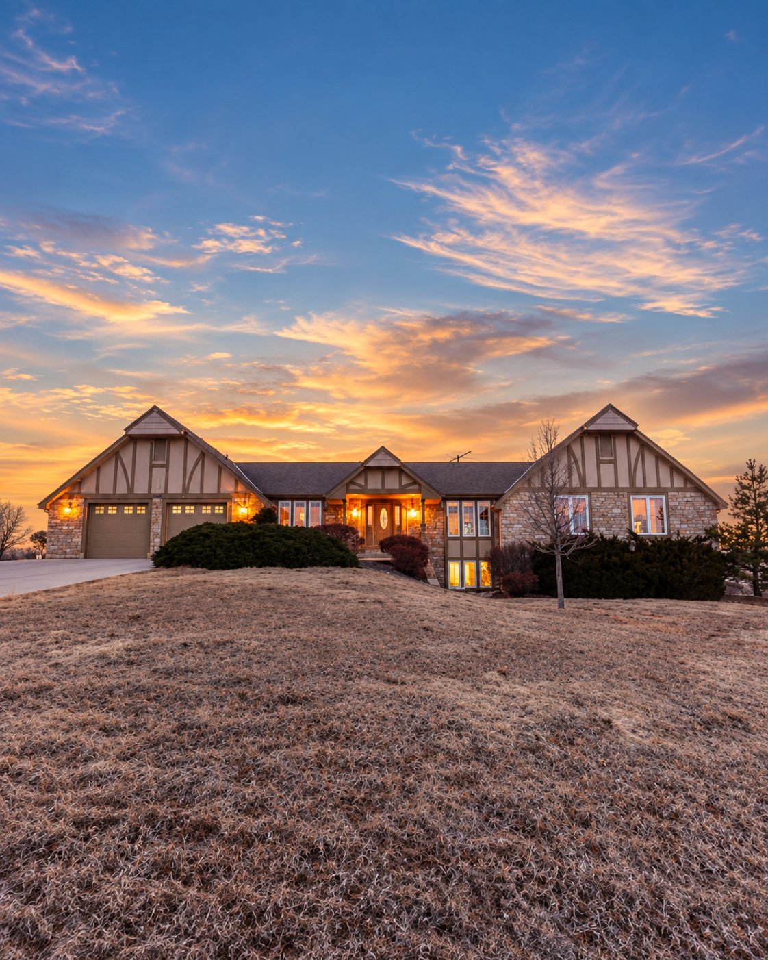 Stone-and-timber house at sunset, with a lit entry and broad dormant lawn.