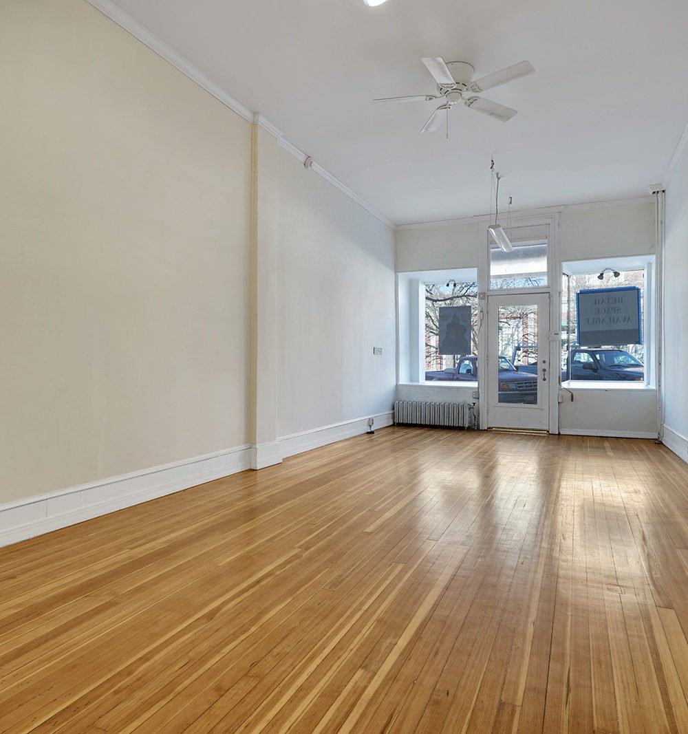 Vacant retail storefront with polished hardwood floors and a glass entry door flanked by large windows.