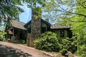 Rustic home with a stone chimney, covered porch, and curving driveway beneath mature trees.