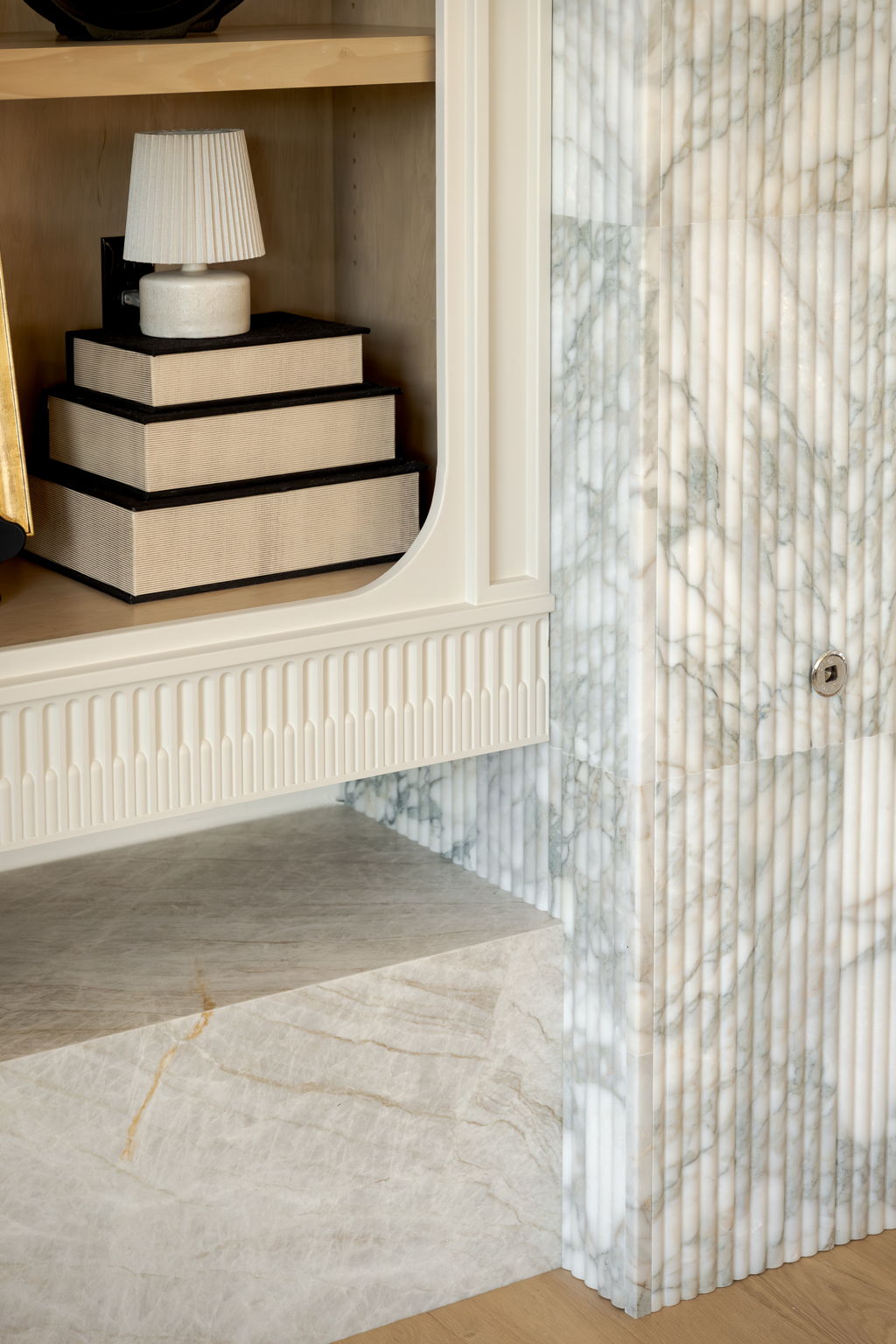 Fluted marble partition beside a recessed niche with stacked boxes and a lamp.