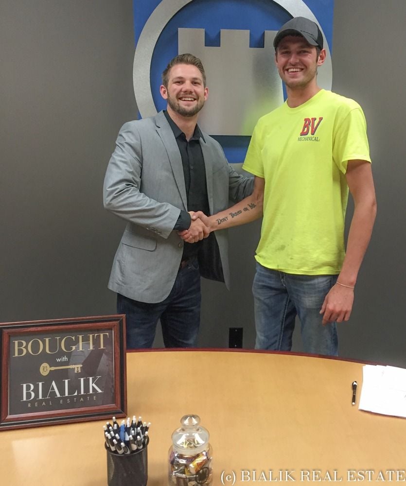Two men shake hands at a real estate closing beside a framed Bialik Real Estate sign.