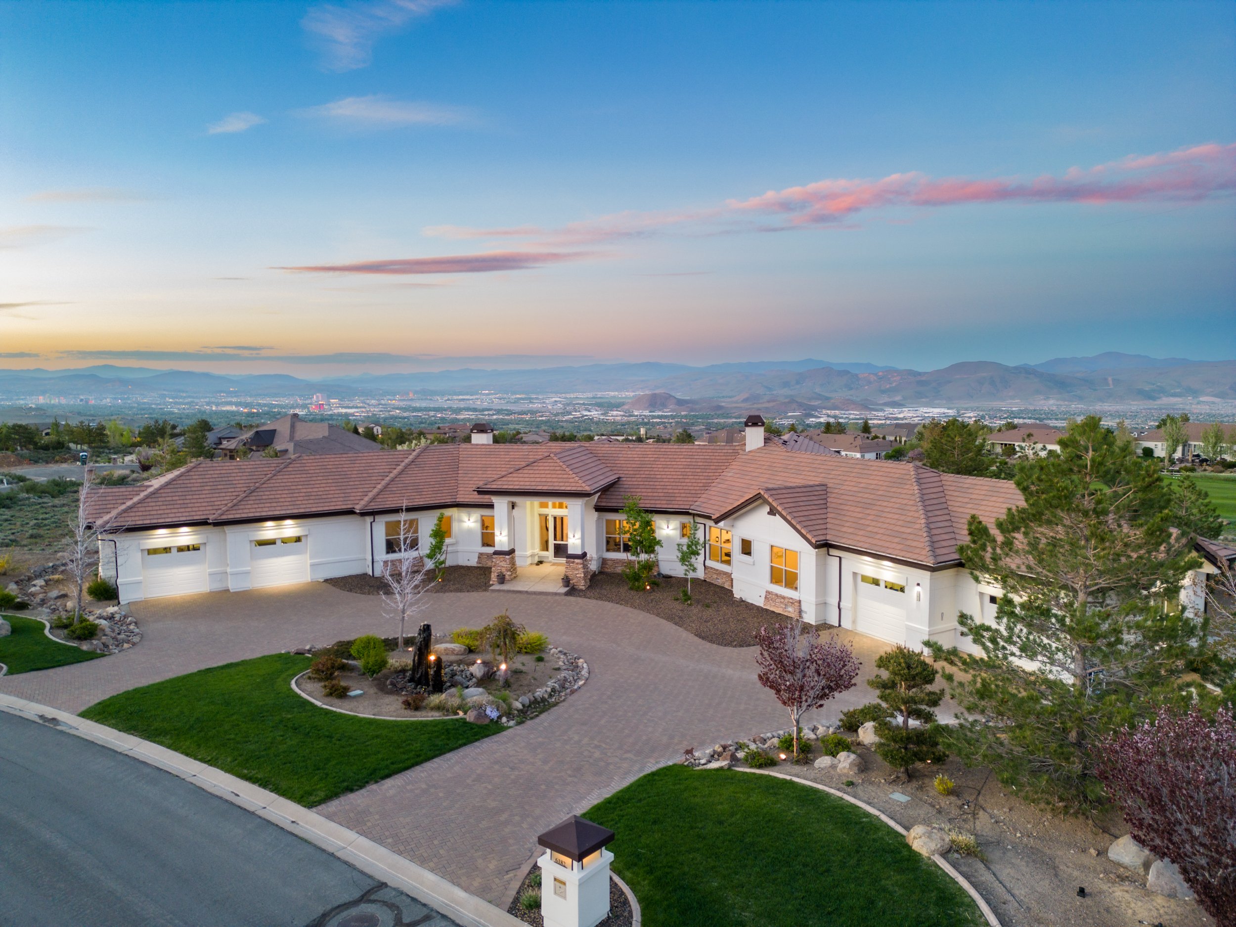 White single-story home with a curved driveway, landscaped grounds, and distant mountain views at sunset.