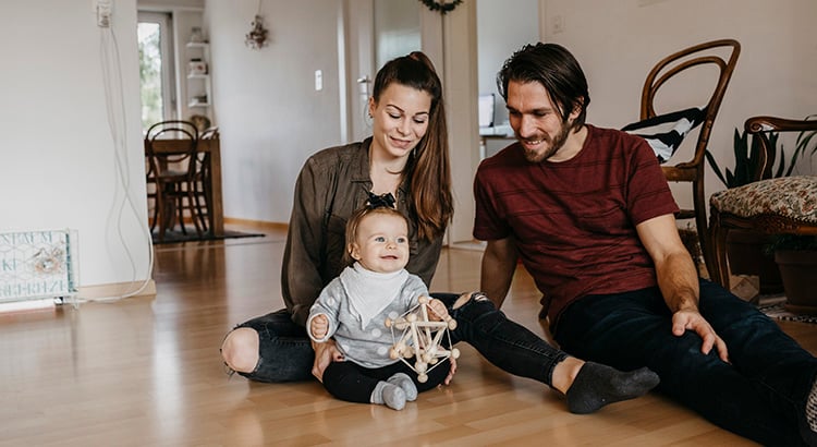 A couple sits on a hardwood floor with their baby and a wooden toy.