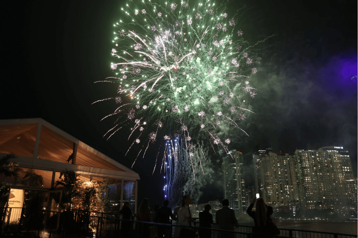 Fireworks burst above a waterfront hotel and high-rises as people watch from a terrace.