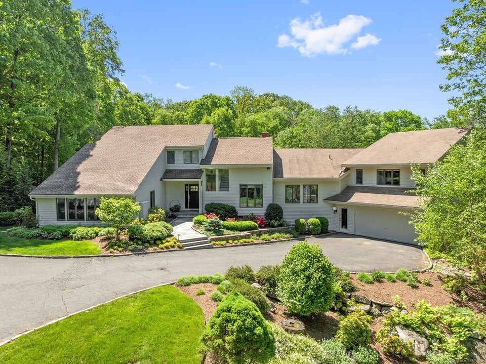 Large light-colored house with an attached garage, curved driveway, and landscaped gardens among mature trees.