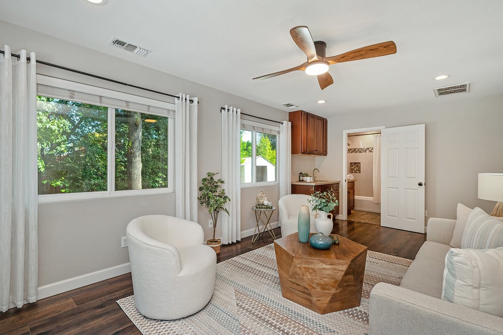 Living room with large tree-facing windows, a wood ceiling fan, and a small adjoining kitchenette.