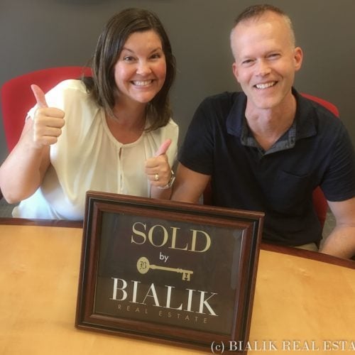 Two smiling adults give thumbs-up behind a framed SOLD sign.