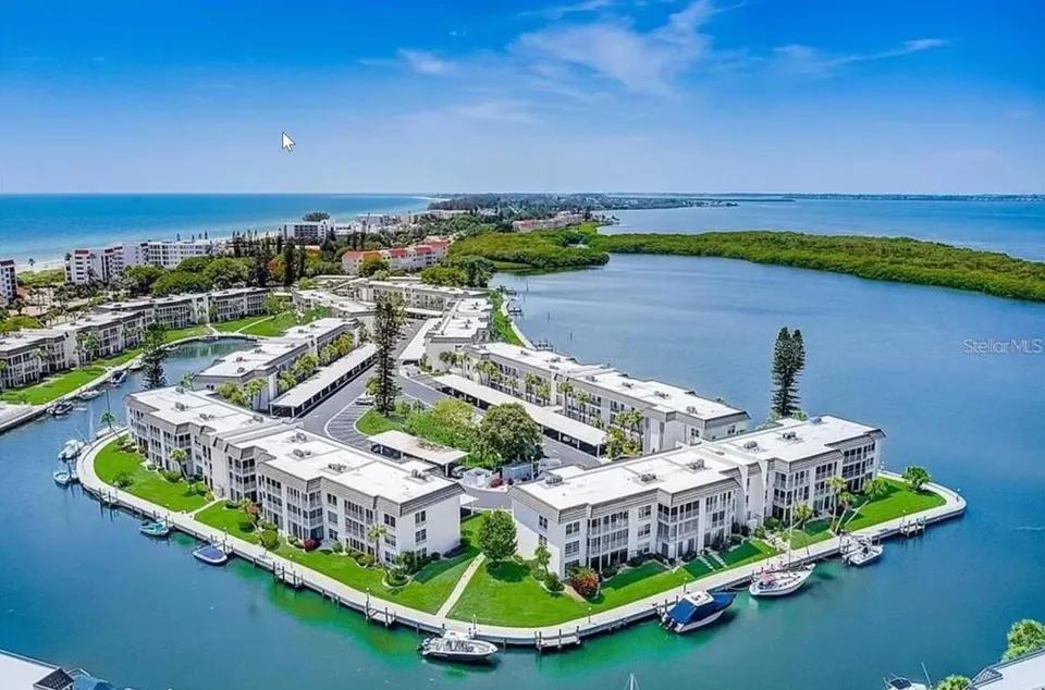 Aerial view of waterfront condominiums with docks and boats between a bay and the Gulf shoreline.