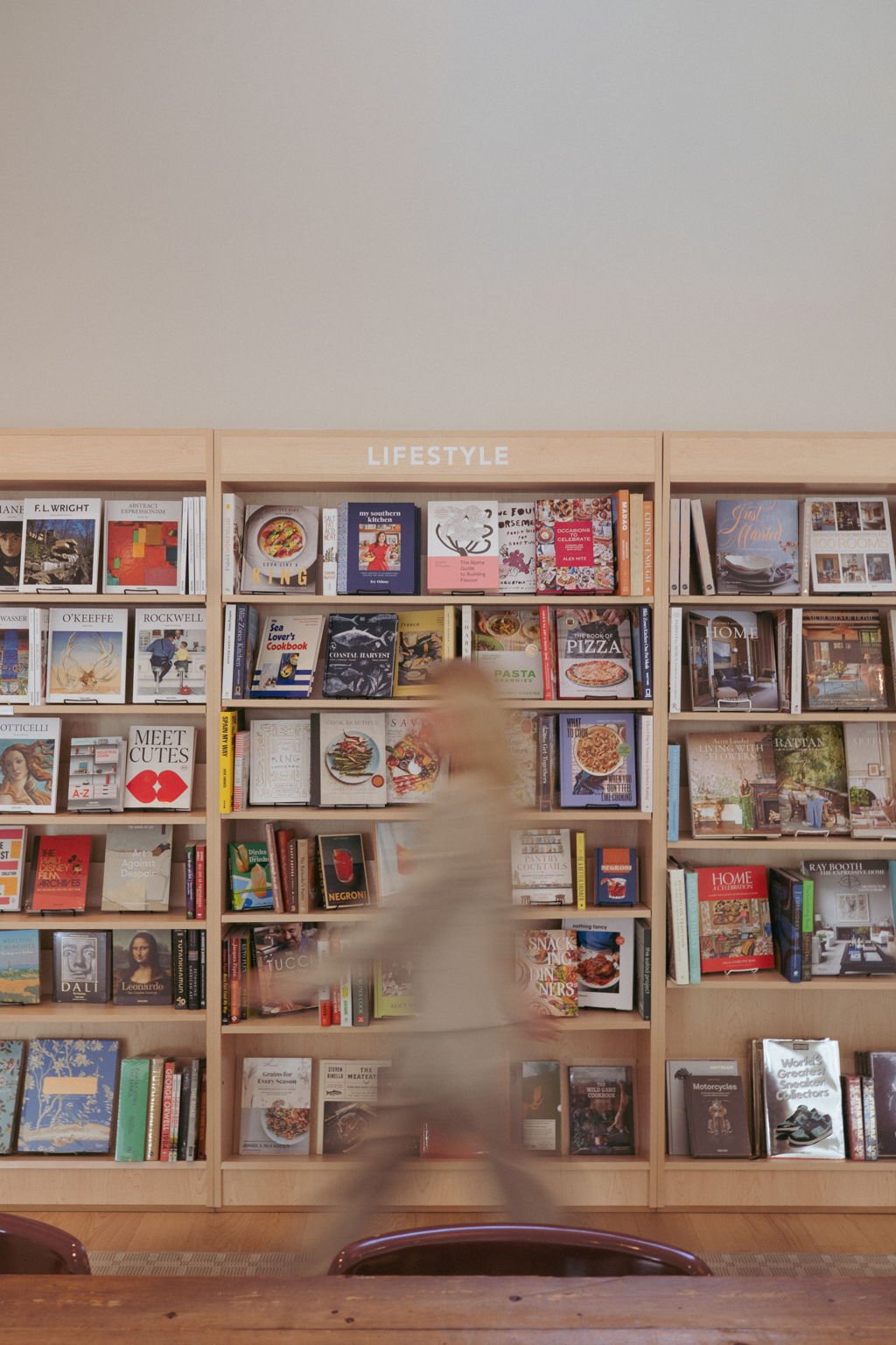 A blurred person browses wooden bookshelves filled with cookbooks, art books, and home design books.