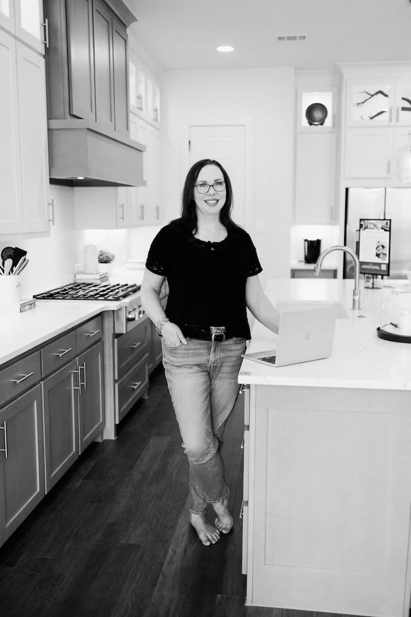A smiling woman in glasses stands barefoot beside a kitchen island with a laptop.
