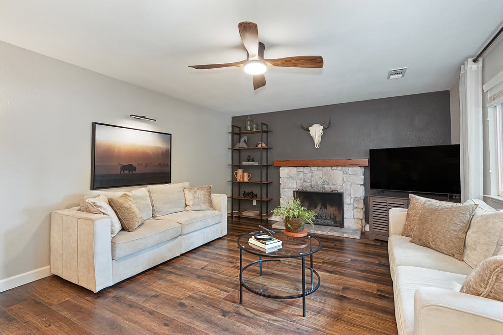 Living room with cream sofas, a stone fireplace, dark wood floors, and a ceiling fan.