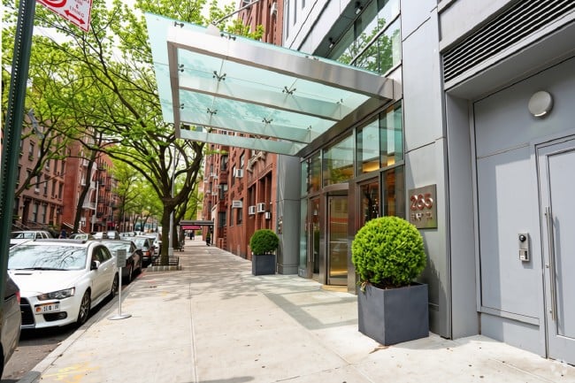 Building entrance beneath a glass canopy, with potted shrubs and a tree-lined sidewalk.