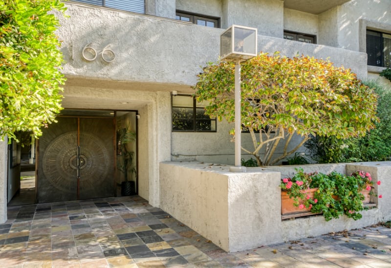 Apartment building entrance with patterned double doors, tiled walkway, and flowering planters.